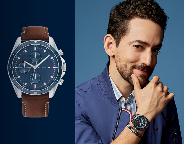 Imagen de un hombre tocando su rostro con la mano derecha dejando ver el reloj que usa de la marca TOMMY HILFIGER. La otra mitad de la imagen está ocupada por el mismo reloj que usa el modelo, donde se puede observar el detalle de la carátula azul y correa café. Relojes Hombre